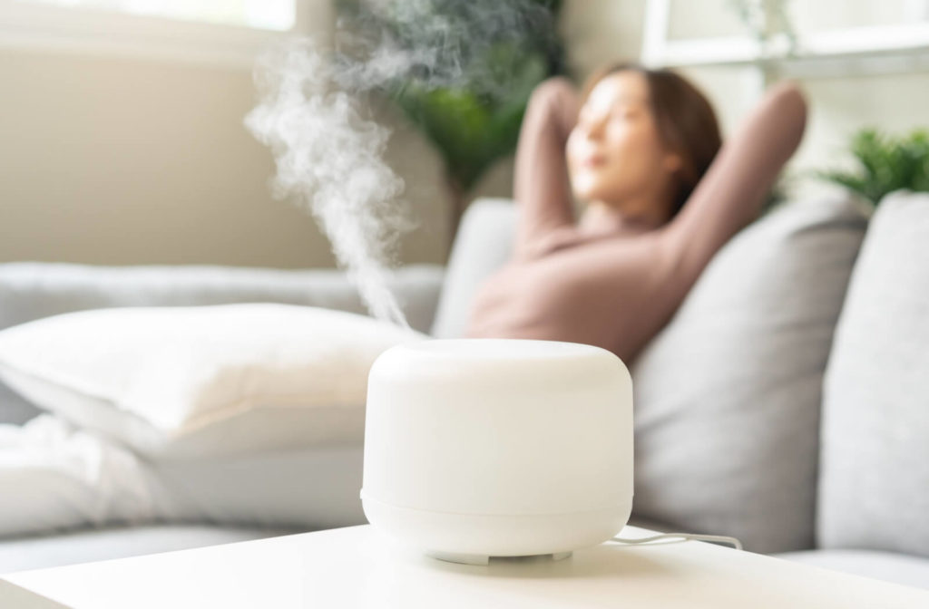 A close-up of a humidifier being used in the living room while a young woman is resting on the sofa.