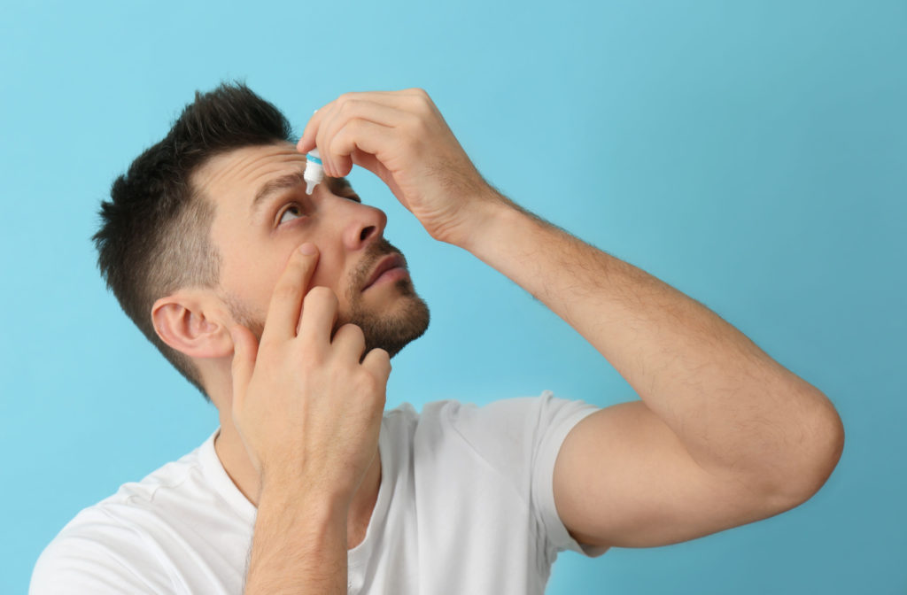 A young man standing against a blue background and looking up as he applies eye drops to his right eye.