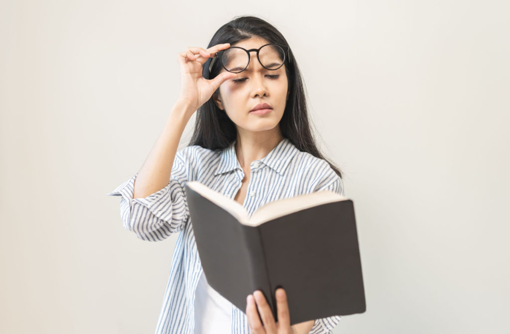 A farsighted woman in a striped shirt holding a book at arm's length, and lifting her glasses in order to read the text clearly.