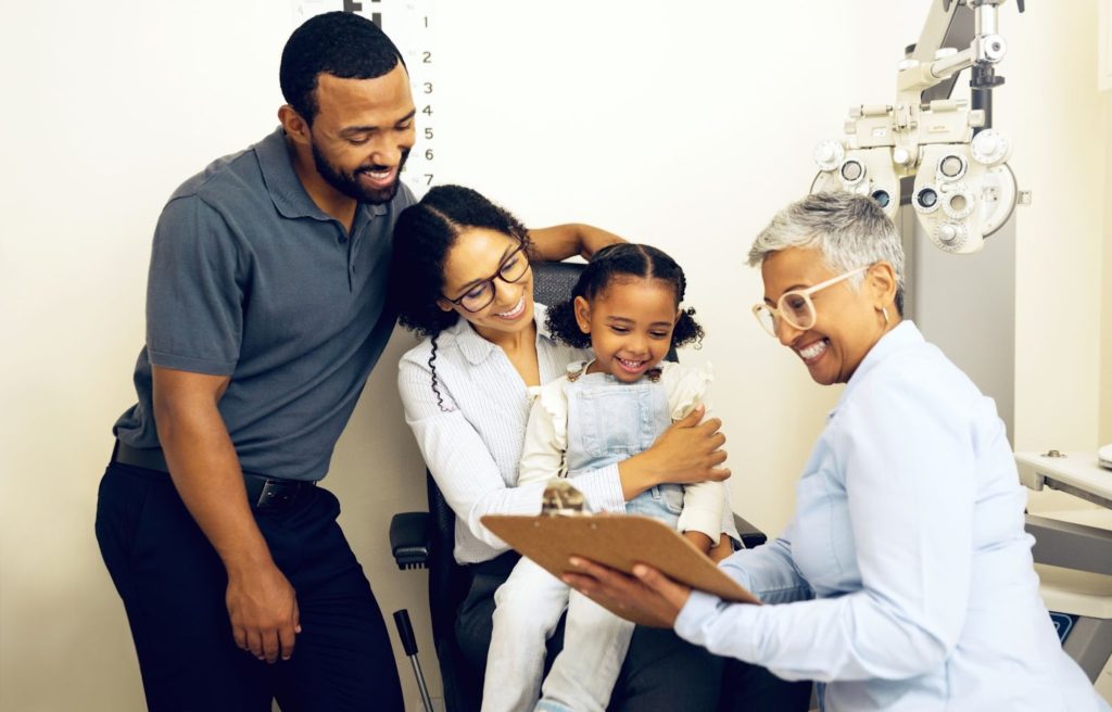 A mother and father sit with a young girl at her first eye exam, smiling with the eye doctor as they review her results.