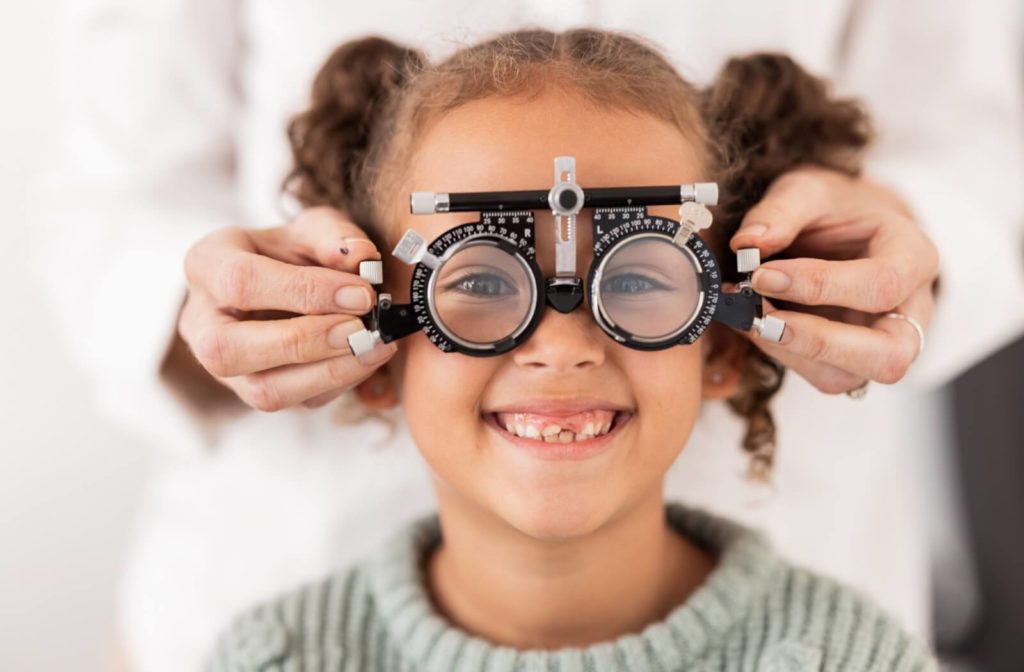 A smiling child looks through lenses being held in front of her eyes by an optometrist.