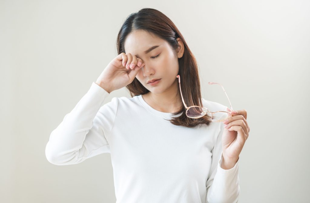 young woman with white long-sleeves rubbing tired eyes for relief while holding a pair of glasses with other hand