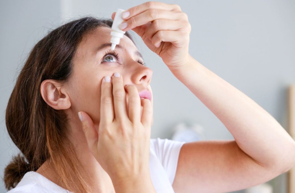 woman with white shirt and brown hair applying eye drops on one eye