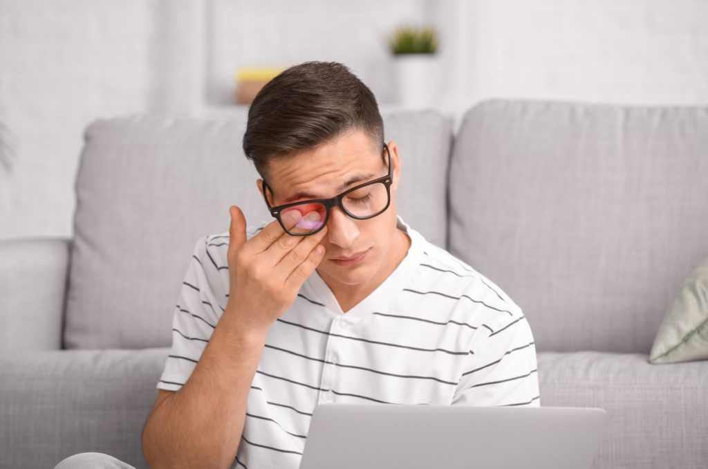 A man with glasses sitting in front of a laptop, rubbing his right eye with his right hand due to dry eyes.