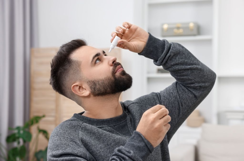 A man looking up while holding a bottle of eye drops in his left hand and over his left eye.