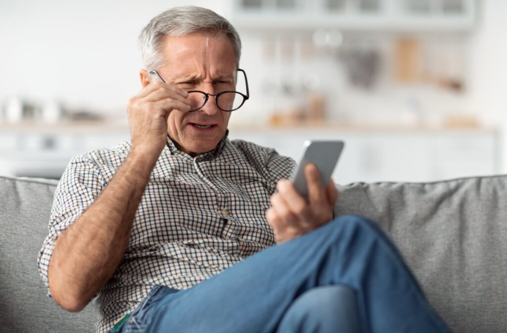 An older patient sitting on their couch, lowering their glasses and squinting at their phone in frustration.