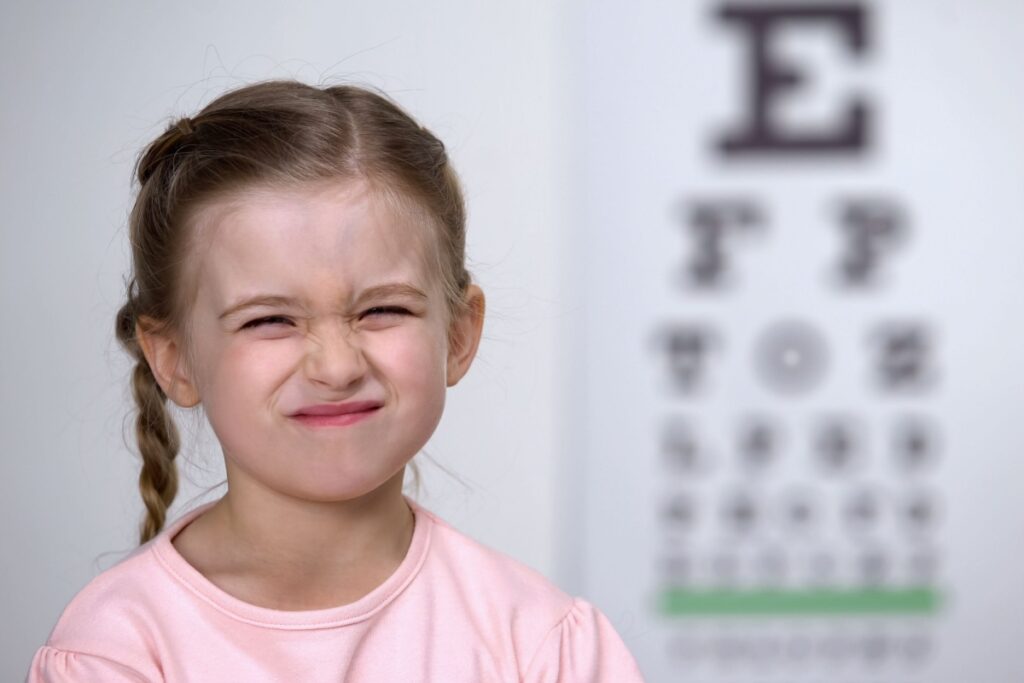 Young girl in pink shirt squinting with strained expression during vision test with blurred eye chart in background.